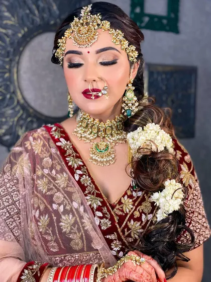 A beautiful bridal portrait with a focus on the complete look. The bride wears a maroon lehenga with heavy jewelry, and her makeup features a bold red lip and shimmering eyes, perfect for her wedding day.