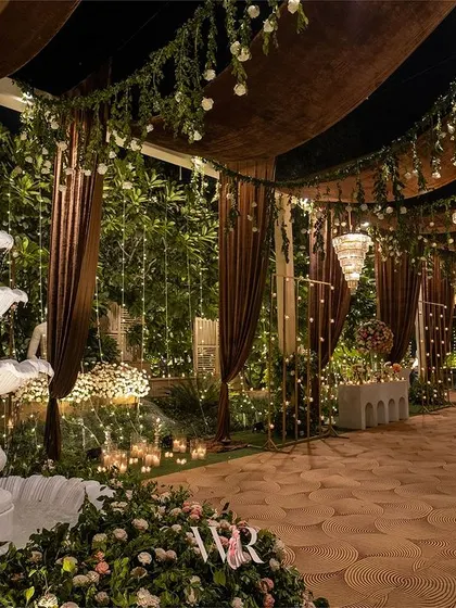 The entrance to a dreamy reception. A serene white fountain surrounded by flowers and candlelight welcomes guests into a space defined by rich brown drapes, fairy lights, and the promise of an elegant evening.