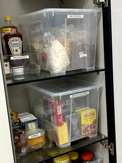 This pantry uses large, clear bins with labels to store backstock items like dals, pulses, and canned goods. This system keeps the pantry tidy and makes grocery planning simple.