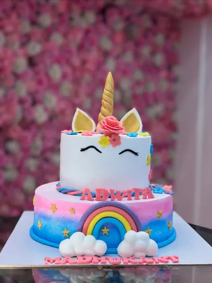 A two-tiered unicorn cake with a rainbow on the bottom tier and a cute unicorn face on top. The fondant clouds and stars complete this enchanting design.
