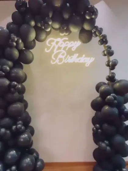 A minimalist yet bold all-black balloon arch. The 'Happy Birthday' neon sign provides a striking contrast against the dark, glossy balloons.