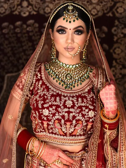 A beautiful bride wearing our rental red velvet lehenga with peacock embroidery. The look is accessorized with a striking green-beaded Kundan necklace and matching maang tikka.