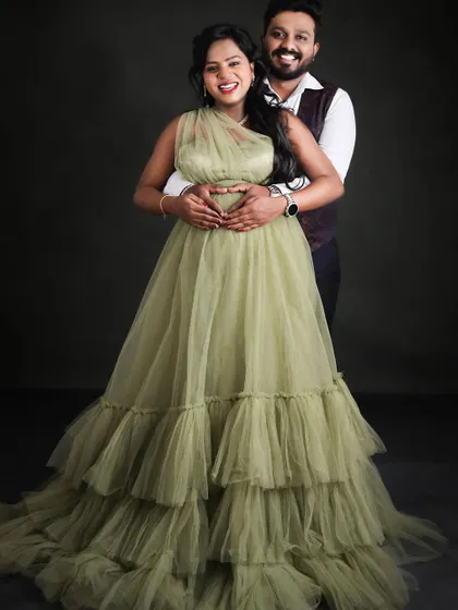 A joyful couple's portrait against a dark studio backdrop. The partner embraces the mom-to-be from behind, both sharing a happy moment.