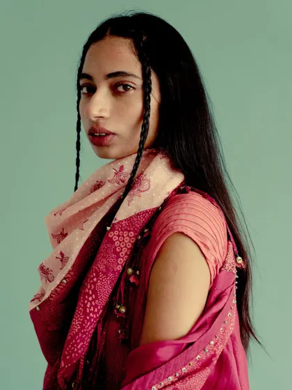 A portrait for Swatti Kapoor against a solid teal background. The warm tones of the pink and red outfit pop against the cool backdrop, highlighting the model's braided hair.