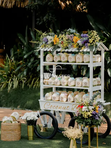 A charming white cart serves as a coconut water station, adorned with colorful flowers to match the event's vibrant theme.