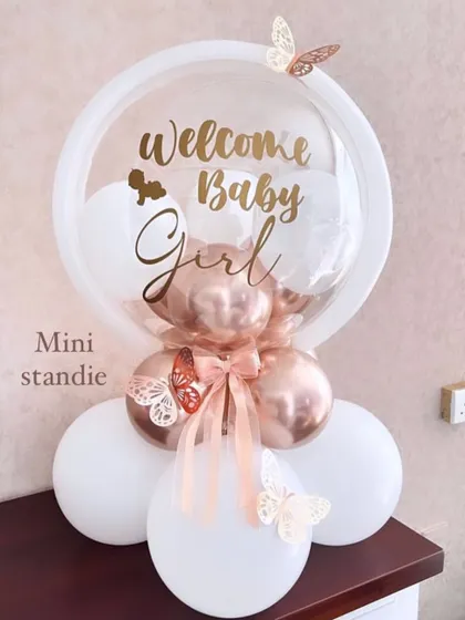 A sweet and simple balloon standee to welcome a baby girl. This arrangement features a personalized "Welcome Baby Girl" bubble balloon on a base of white and rose gold balloons, accented with paper butterflies.