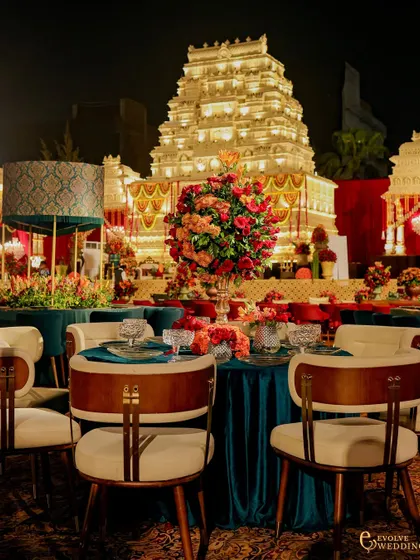 A view of the guest dining tables set against the illuminated mandap. The jewel-toned teal velvet and modern chairs create a stylish contrast with the traditional architecture, showcasing a fusion of styles.