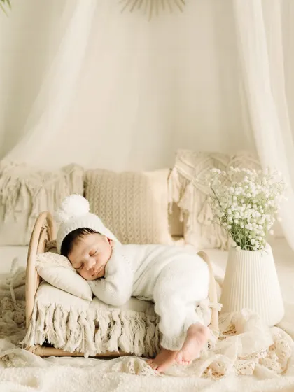 Dressed in a cozy white knit outfit with a pom-pom hat, this newborn sleeps on a miniature wooden bed. The neutral tones and textures create a serene and timeless look.