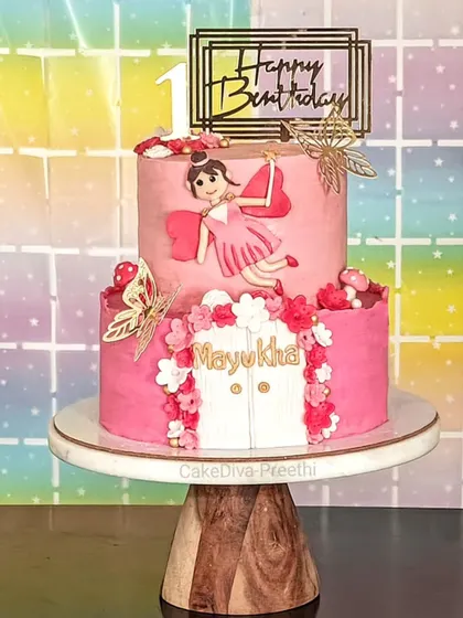 A magical two-tier fairy cake for a first birthday. It features a fondant fairy, a whimsical door, and delicate flowers, all in a lovely pink palette to make the celebration extra special.