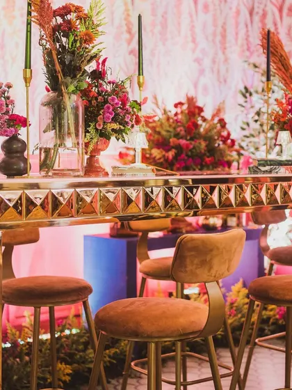 A contemporary sangeet bar setup, featuring a gold-studded bar table, velvet stools, and vibrant red floral arrangements against a pink patterned backdrop.