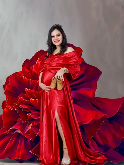 A stunning and creative studio portrait. The mother-to-be holds a pair of baby shoes while wearing a vibrant red gown that transforms into a giant floral bloom.