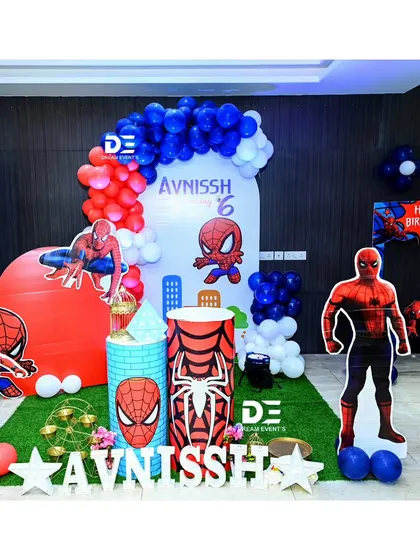 A cool Spiderman setup for Avnissh's 6th birthday. This decor includes a cityscape backdrop, multiple Spiderman cutouts in different poses, and a red, white, and blue balloon arch.