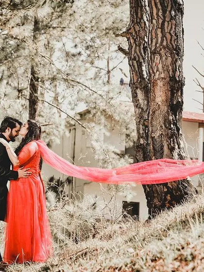 A dynamic and romantic pre-wedding photo in a forest setting. The bride's vibrant red dupatta flows in the wind, creating a beautiful sense of movement as the couple shares an intimate moment.