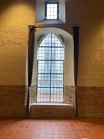 A magnificent arched window inside the Belfort Museum in Bruges. The scale of the window, combined with the exposed brick wall and terracotta floor, creates a powerful sense of history and space.