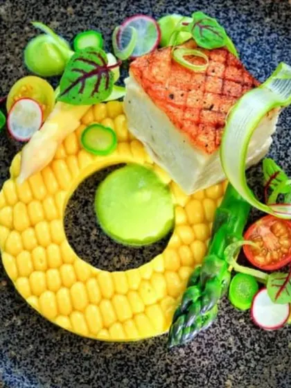 A creative dish featuring a corn-shaped element with seared fish and fresh vegetables. Our chefs love to play with shapes and ingredients to surprise and delight your guests.