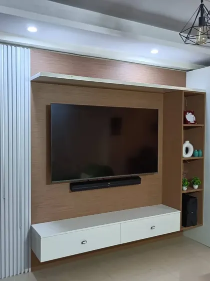 This living room features a custom TV unit with an integrated pooja mandir. The design cleverly combines entertainment and spirituality with white vertical paneling and light wood finishes.