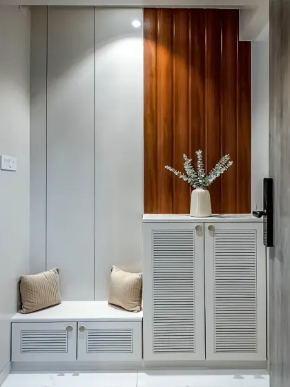 A minimalist entryway design with a white seating bench and a matching shoe cabinet with louvered doors. The dark wood accent panel adds warmth and texture to the clean, white space.