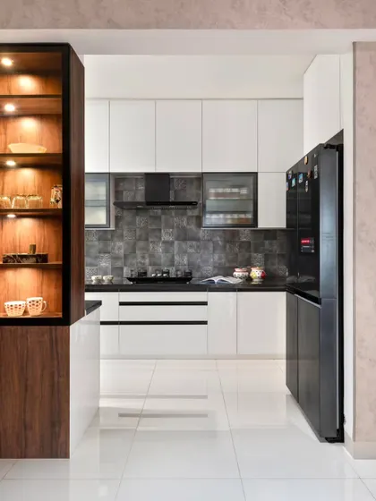 This modern kitchen in the 'Muditā' project features a striking contrast of high-gloss white acrylic cabinets and a dark, textured Spanish tile backsplash. A bar unit provides a visual break from the living area.