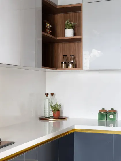 In this kitchen, a familiar fusion of white, grey, and blue exudes a modern yet classic charm. An open corner shelf in a wood finish breaks the monotony of the glossy cabinets.