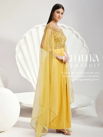 The Emma Jumpsuit in a vibrant yellow. This unique piece features an embellished off-shoulder bodice, dramatic cape sleeves, and wide-leg pleated trousers for a modern and graceful statement.