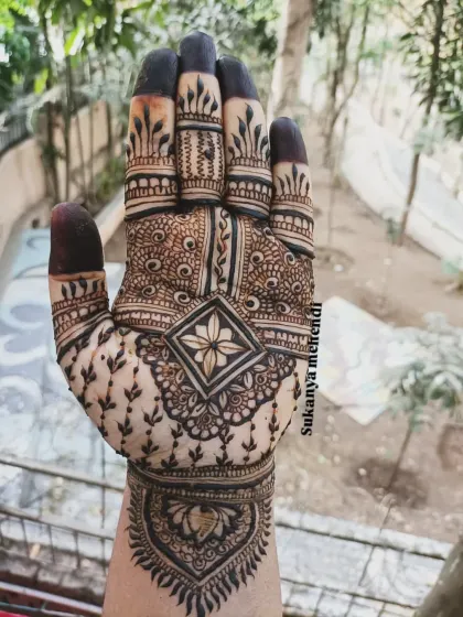 A close-up of a detailed palm design. The central floral element is surrounded by delicate vines and geometric patterns, with bold henna on the fingertips.