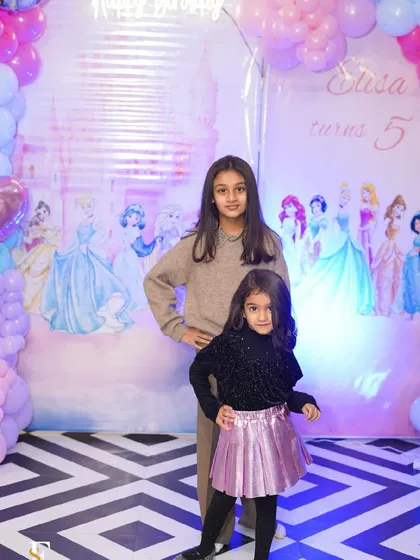 Two sisters pose in front of a Disney princess backdrop, surrounded by beautiful pink and purple balloons.