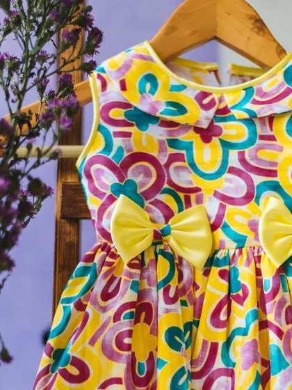 A vibrant and colorful abstract print dress in shades of yellow, purple, and teal, adorned with two cheerful yellow bows.