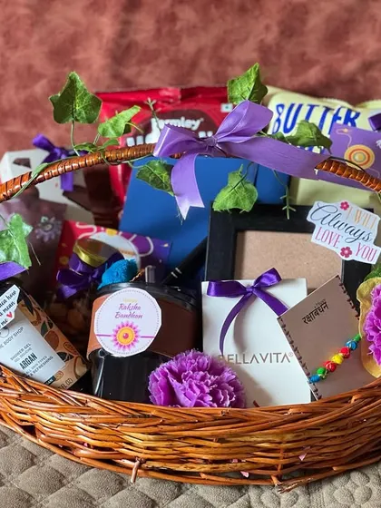 A wicker basket Rakhi hamper filled with goodies and decorated with purple flowers and ribbons, creating a vibrant and cheerful gift.