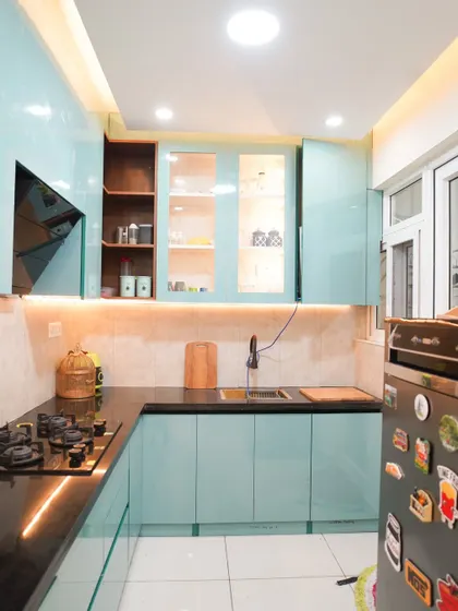 A bright and cheerful L-shaped kitchen with glossy aqua-blue cabinets and a black granite countertop. The design includes glass-fronted upper cabinets with internal lighting to display glassware.