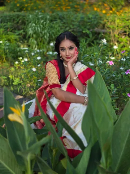 The vibrant red and white of the Bengali saree pop against the lush greenery. This pose, with her hand gently touching her face, adds a touch of classic elegance to the portrait.