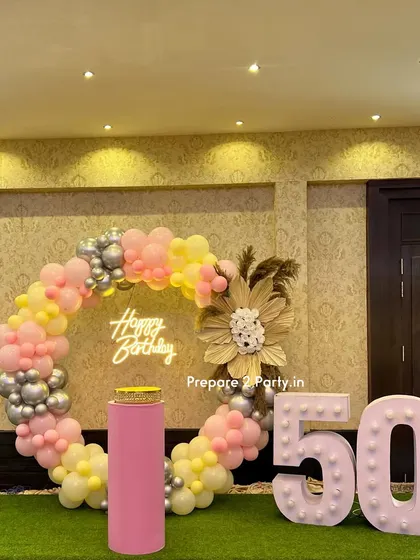 A cheerful and stylish 50th birthday setup with a circular balloon arch in pastel colors. The design is accented with a dried flower arrangement, a neon sign, and large marquee numbers on an artificial grass base.