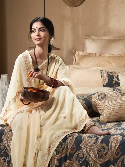 This image for the 'Sanskriti' collection contrasts the light, traditional saree with the dark, rich bedding. The composition is elegant and tells a story of quiet moments at home.