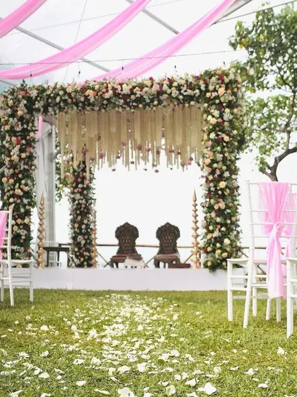 An open-air mandap under a clear tent, decorated with pink drapes and lush florals for an intimate garden wedding feel.