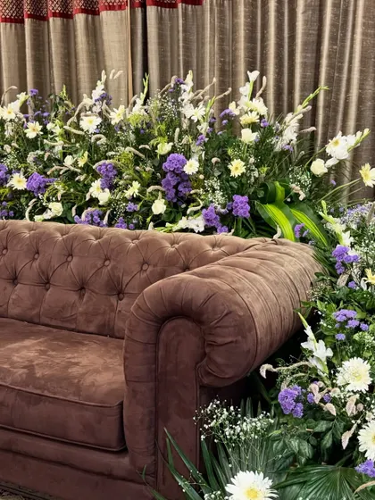 A luxurious floral arrangement featuring white lilies and purple statice, designed to sit behind a classic tufted brown sofa.