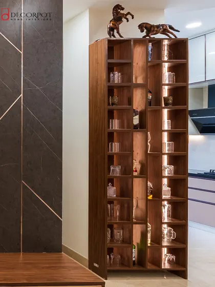 A tall, elegant wooden shelving unit used as a bar or display case. The asymmetrical design and internal lighting make it a beautiful statement piece for a living or dining area.