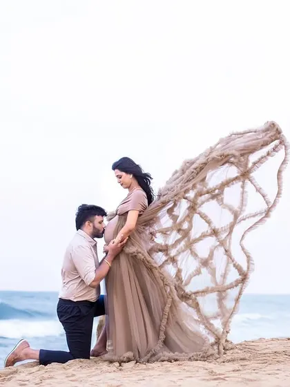 A tender moment between the couple, with the father-to-be kissing the bump. The unique gown adds a touch of fantasy to this beach scene.