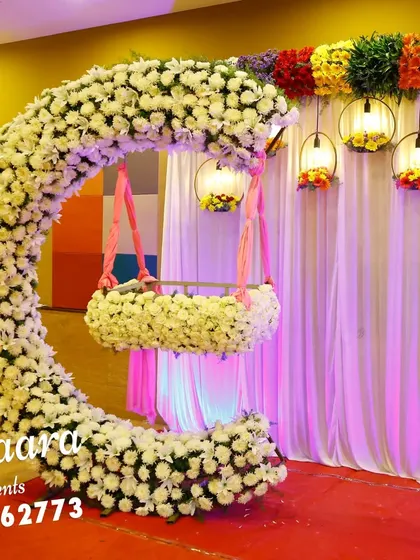 A stunning cradle ceremony setup featuring a crescent-shaped arch made of white flowers. The hanging cradle, also adorned with flowers, creates a beautiful focal point, while colorful lanterns add a festive touch.