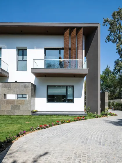The curved driveway at Triveni Farmhouse softens the approach to the home's geometric facade. Vertical wooden louvers on the balcony add a touch of warmth and provide privacy while contributing to the building's modern aesthetic.