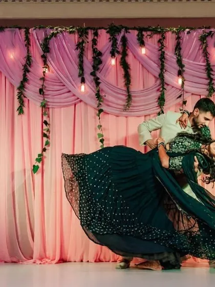 A dramatic and romantic dance move captured during a Sangeet performance, with a backdrop of soft pink drapes and hanging lights.