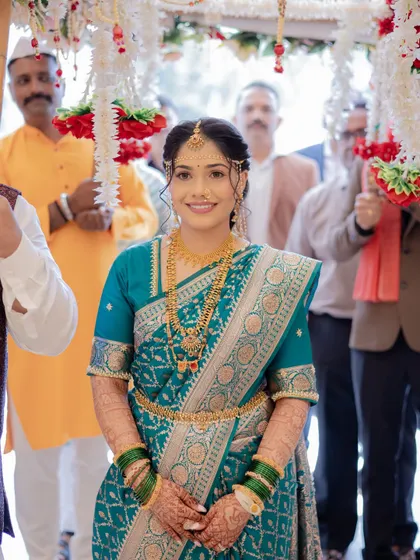 The bride's grand entrance. This shot captures her walking under a floral canopy, looking serene and beautiful in her teal blue wedding saree and traditional gold jewelry.