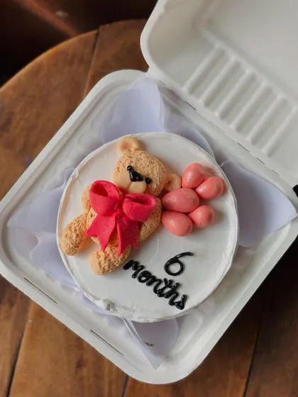 An incredibly cute bento cake for a 6-month-old's celebration. It features a hand-piped teddy bear with a big pink bow, holding a bunch of pink balloons.