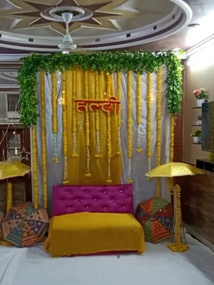 An indoor Haldi seating area with a backdrop of white and yellow drapes, a green foliage border, and hanging marigold strings. The setup is completed with decorative umbrellas.