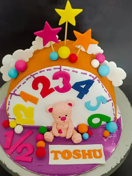 A colourful half-birthday cake with a clock theme, a cute teddy bear, and bright numbers.