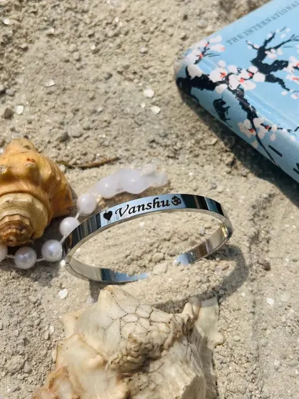 This silver Kada, engraved with 'Vanshu' and symbols, is captured in a beachy setting with a book and pearls, giving it a serene and personal feel.