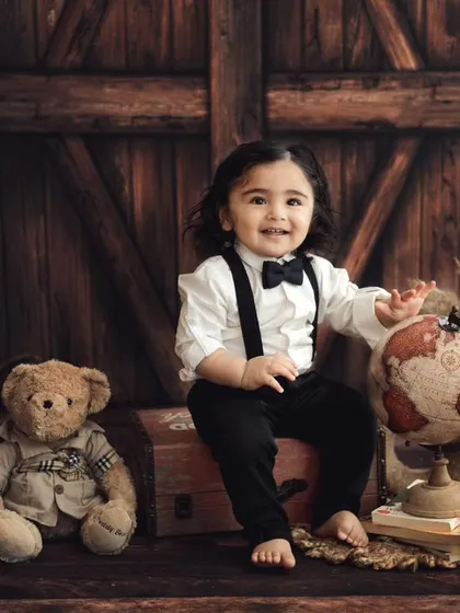A dapper little traveler ready for his first birthday adventure. Dressed in a bow tie and suspenders, he's sitting on vintage-style trunks next to his teddy bear companion.