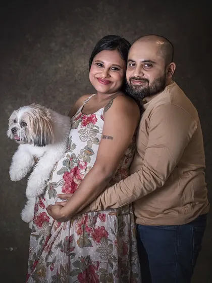 An expecting mother holds her dog close to her baby bump, with her partner embracing her from behind. It's a sweet photo that shows the whole family, including the furry members, sharing the moment.