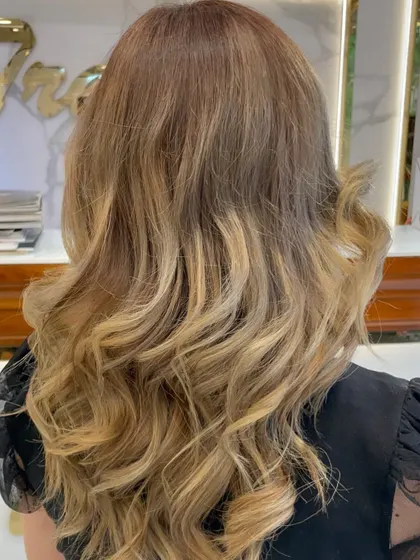 Golden hour for your hair. This sun-kissed balayage blends darker roots into bright, golden blonde ends, styled in loose curls for a perfect beachy vibe.