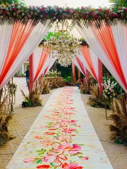A stunning wedding entrance pathway featuring a printed floral carpet, elegant drapes, and chandeliers. The aisle is lined with pampas grass for a modern, bohemian touch.