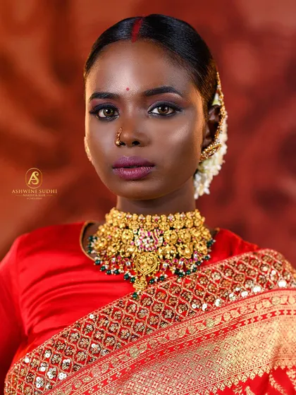 A striking close-up of a bride in a red saree. The focus is on her expressive eyes and the intricate details of her gold choker necklace.