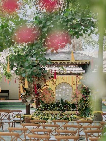 A beautifully framed shot of the Haldi mandap, seen through the lush trees of the venue, creating a sense of a hidden, magical space.